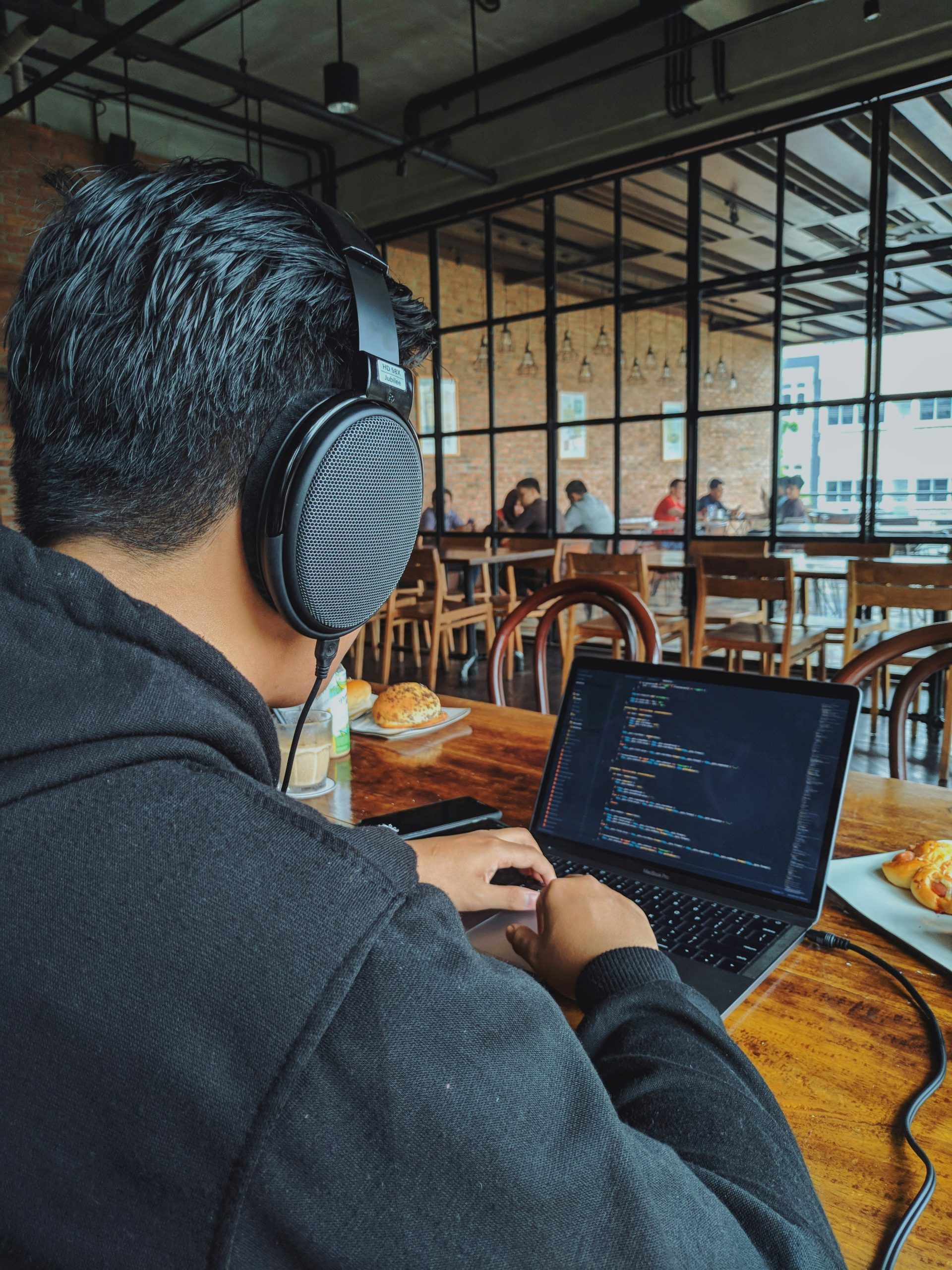 Person wearing headphones working on a laptop in a café, focused on coding, with pastries and drinks on the table, illustrating the importance of technical proficiency and communication skills for executive assistants.